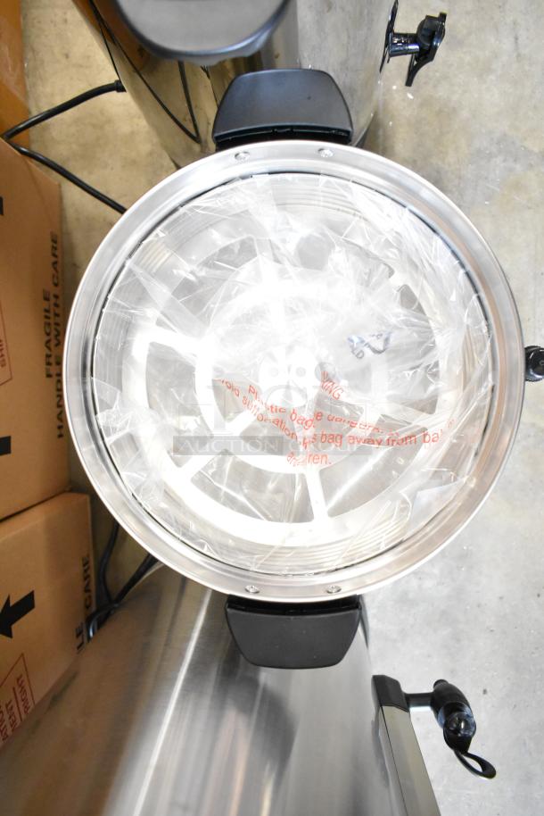 Stainless steel coffee urn with plastic packaging inside, viewed from above. Handle and spout visible. Scratch and dent condition.