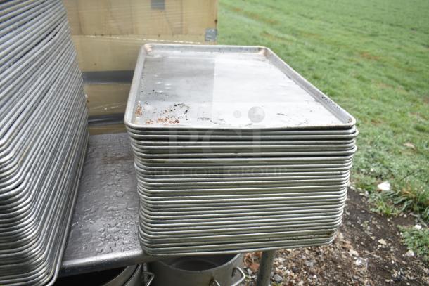 104 full-size metal baking pans stacked outdoors, showing some wear and rust. Auction offers 104 pans per bid.