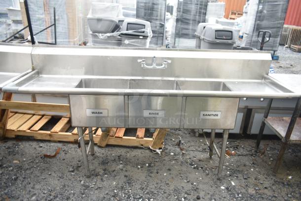 Commercial 3 bay sink with dual drain boards, labeled "Wash," "Rinse," and "Sanitize." Metal construction, outdoor setting.