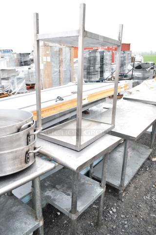 Two commercial metal tables with under shelves, outdoor setting, used condition, suitable for industrial use.