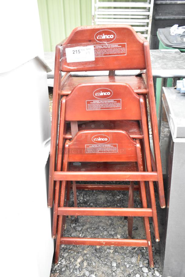 Three Winco wooden high chairs stacked. Sturdy design, natural wood finish, safety harness, some wear visible.