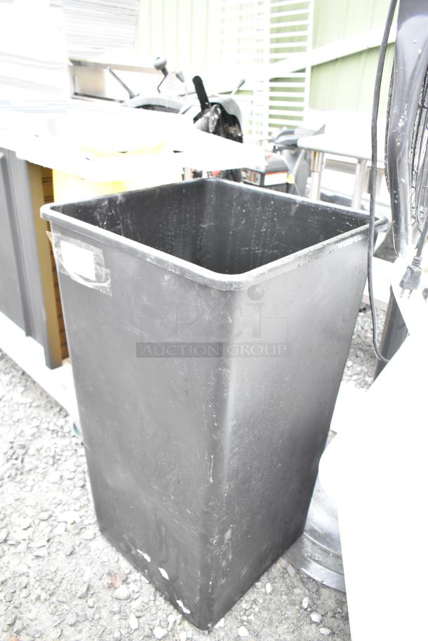 Black poly trash can, rectangular, lightly worn condition, visible tape mark on side, outdoor setting.