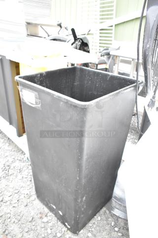 Black poly trash can, rectangular, lightly worn condition, visible tape mark on side, outdoor setting.