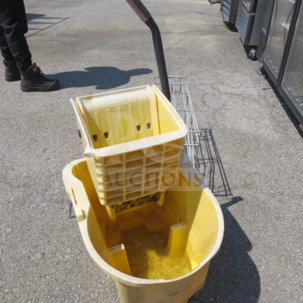 Rubbermaid yellow mop bucket with wringer, used condition, sturdy handle, visible water residue.