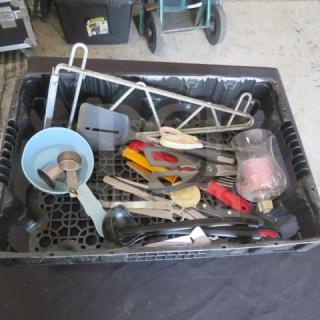 Plastic crate filled with miscellaneous tools and kitchen items, including measuring cups, tongs, scissors, and a ladle. Items are used.
