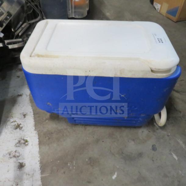 Blue Igloo cooler with white lid, showing wear and dirt. Visible wheels for mobility. Brand name embossed on side.