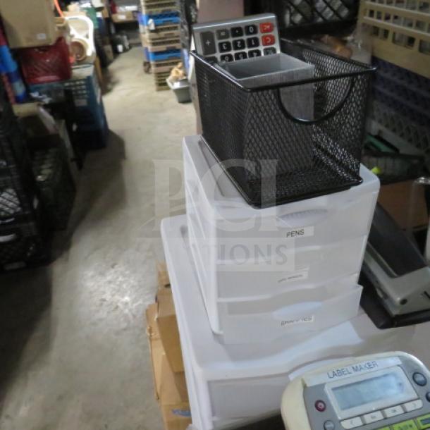 A collection of office supplies including a mesh basket with a calculator, plastic drawer organizers labeled "PENS," a label maker, and assorted items in a storage room.