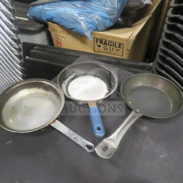 Three used 8-inch saute pans with metal handles on a surface. Blue-handled pan in center, other pans with visible wear.