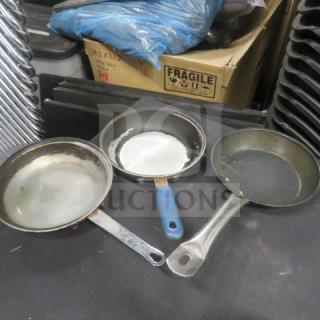 Three used 8-inch saute pans with metal handles on a surface. Blue-handled pan in center, other pans with visible wear.