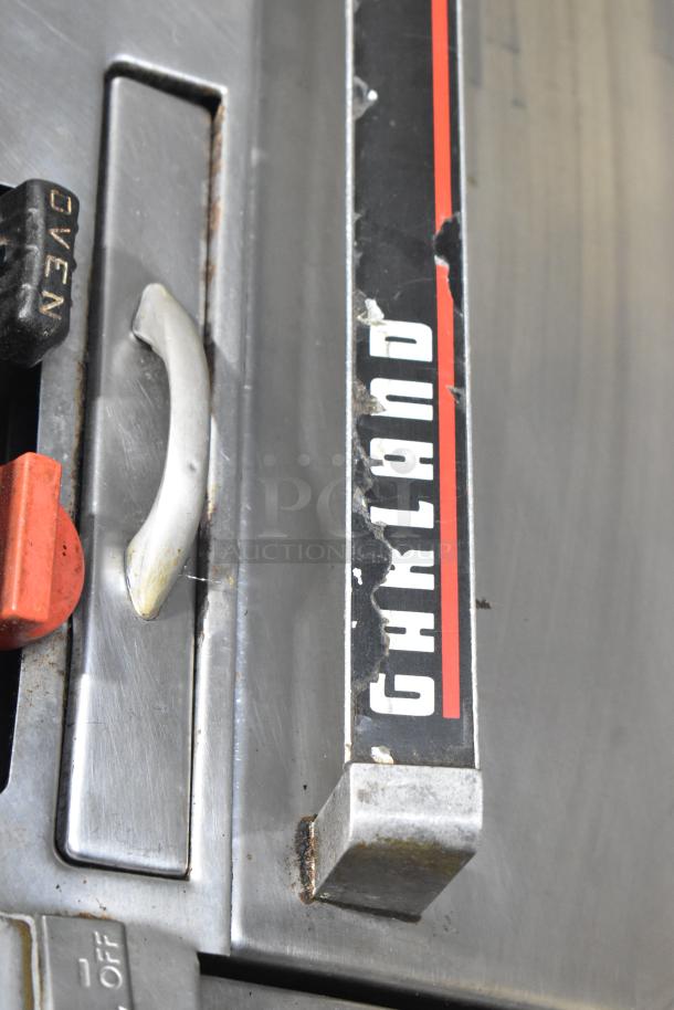 Garland commercial gas-powered 4-burner range with oven, featuring over shelf and backsplash. Visible wear, clear brand marking.