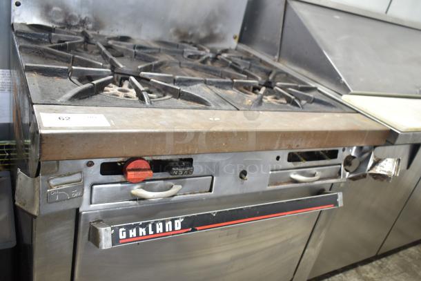 Garland commercial gas-powered 4-burner range with oven, overshelf, and backsplash. Shows wear and tear. Model unidentified.