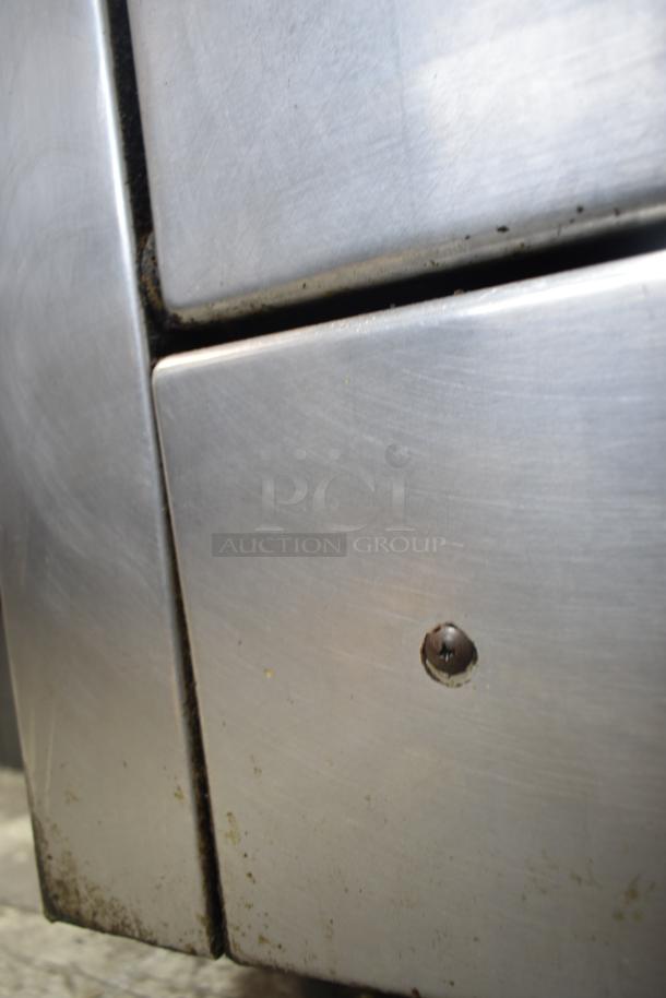 Garland commercial gas range with 4 burners, oven, over shelf, and backsplash. Stainless steel finish, showing signs of wear.