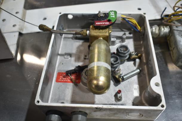 Metal fire suppression box with release mechanism and wiring, visible labels, and components, condition appears used.