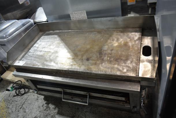 Commercial gas-powered flat top griddle with cheese melter. Stainless steel, well-used condition, visible caution label.
