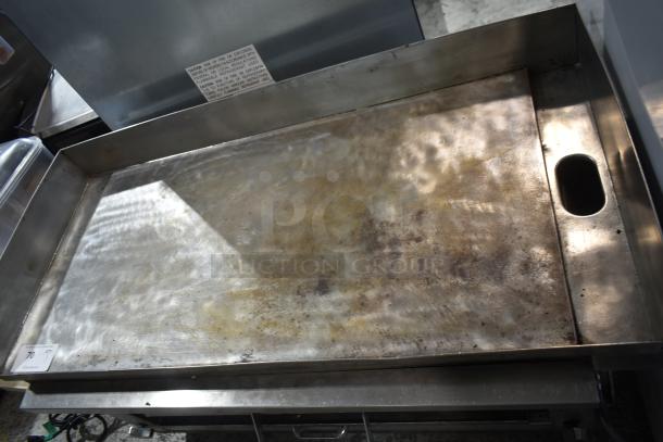 Commercial gas-powered flat top griddle with cheese melter, stainless steel, moderate wear, visible caution label on backsplash.
