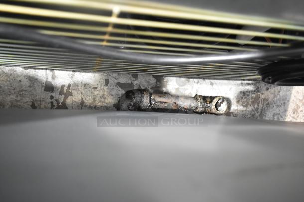 Commercial gas-powered griddle underside view, showing a metal pipe beneath cooking surface. Condition appears to be well-used.