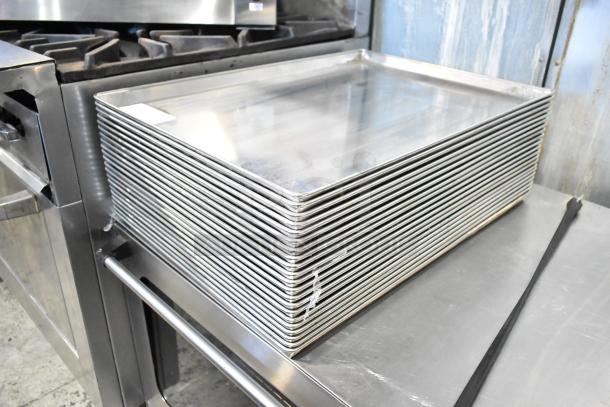 Stack of 21 full-size baking pans, stainless steel, used condition, visible label on top pan, industrial kitchen setting.