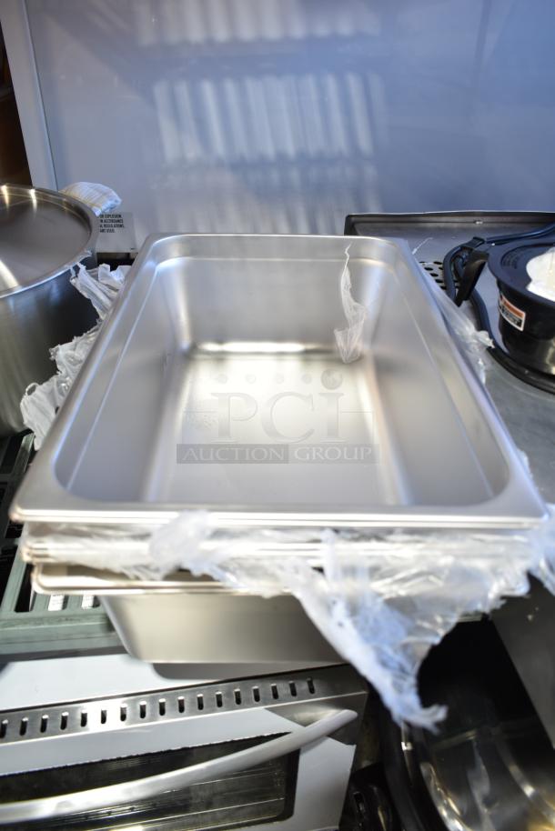 New stainless steel steam table pans, various sizes and brands, stacked, wrapped in plastic. Scratch and dent condition.