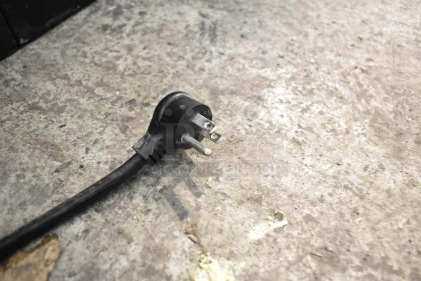Black power plug for 2018 True GDM-49-HC commercial cooler, 115 volts, 1 phase, on a rough concrete surface.