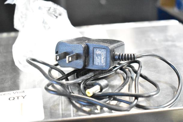 Power adapter for Tor Rey L-PC 40L 40 lb. digital scale. Shows plug and coiled cord on a metal surface.