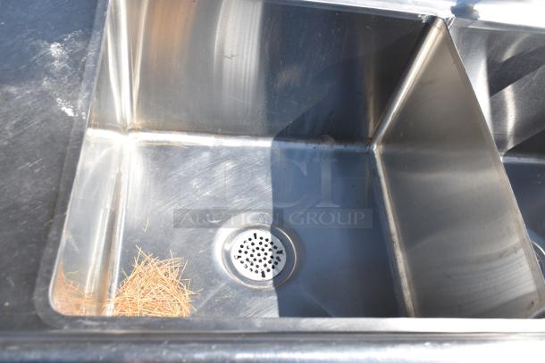 Commercial 3 bay stainless steel sink with dual drain boards, slight wear, visible drain, and leaf debris in one bay.