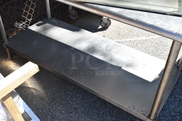 Commercial metal table with under shelf, featuring wheels and a durable stainless steel construction. Visible outdoor wear.