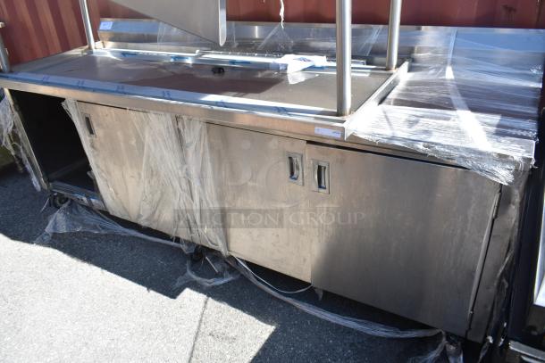 Stainless steel work counter with doors, partially wrapped in plastic, on wheels; includes labeling with unspecified details.
