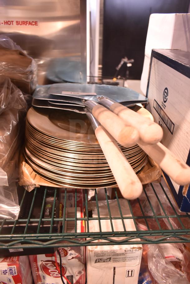 Stack of metal pizza peels with wooden handles on a shelf, alongside a large set of gold plate chargers. 28 items total.