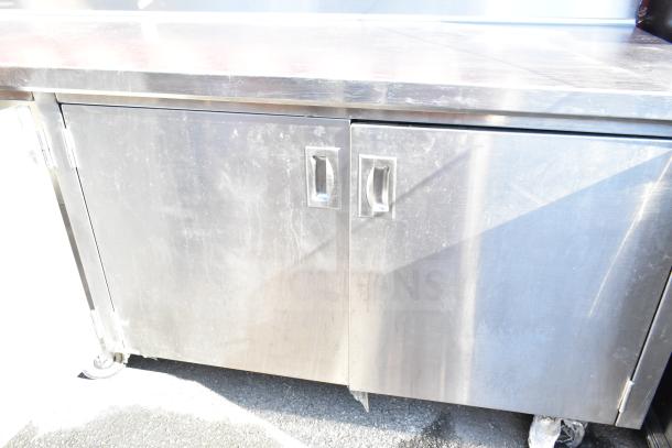 Commercial stainless steel counter with dual doors and under shelf, showing signs of wear. Equipped with casters for mobility.