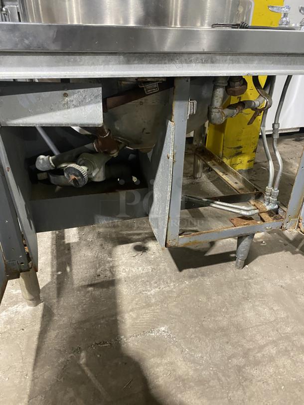 Commercial stainless steel steam kettle with power tilt on legs, showing valves and pipes. Appears in used condition.