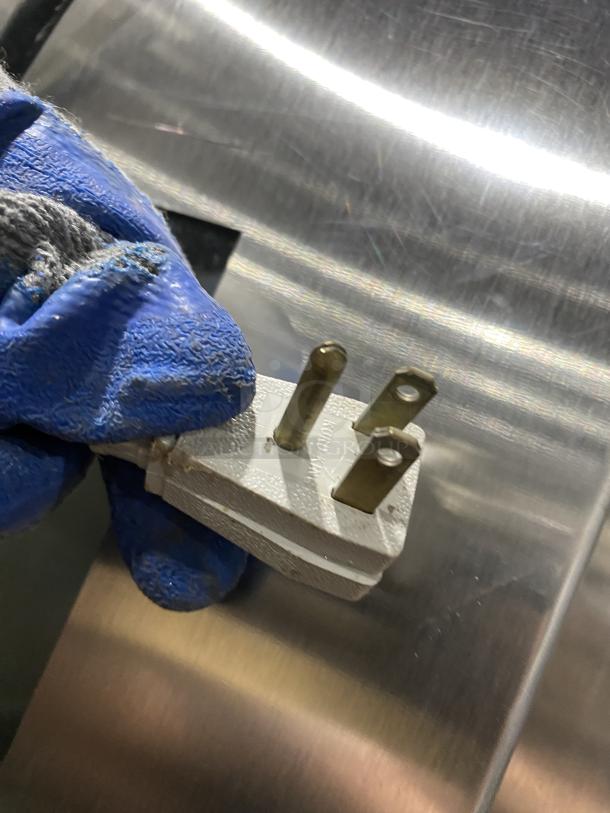 Close-up of a plug held with a gloved hand, against a stainless steel surface, part of a True STG2R-2G refrigerator.