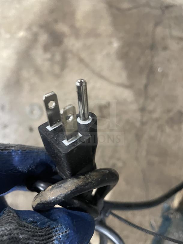 A close-up of a standard three-prong power plug, held in a gloved hand, on a concrete floor.