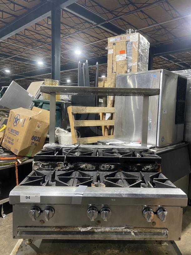 Stainless steel gas-powered 6-burner range on legs, used condition, visible wear on burners, with control knobs and storage beneath.