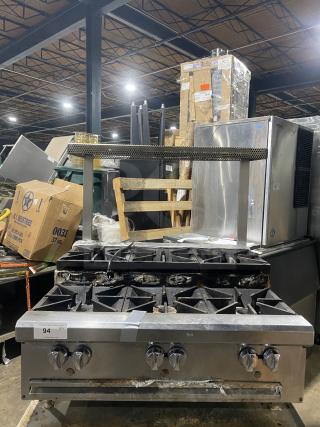 Stainless steel gas-powered 6-burner range on legs, used condition, visible wear on burners, with control knobs and storage beneath.