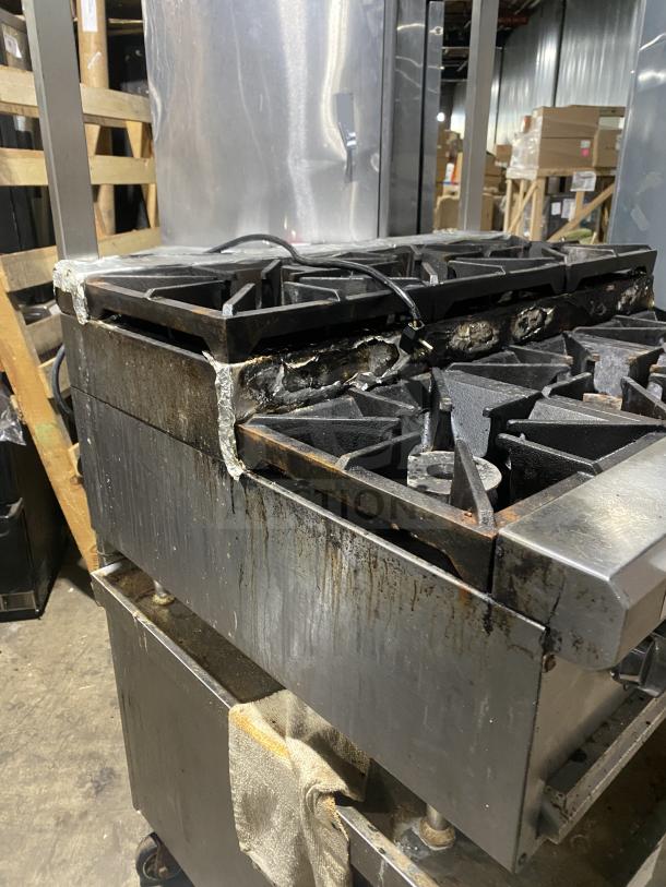 Commercial stainless steel 6-burner gas range on legs. Signs of use and corrosion present; ideal for heavy-duty kitchens.