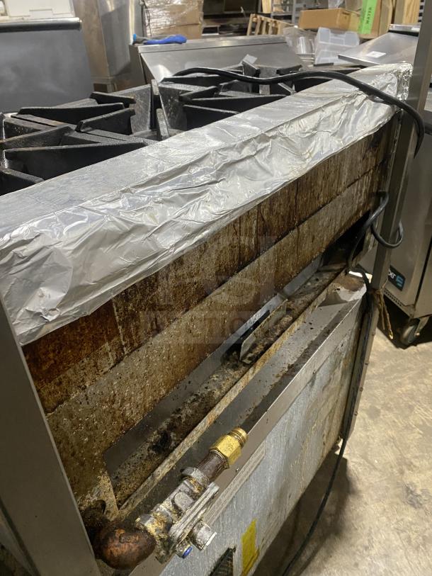 Stainless steel 6-burner gas range on legs, commercial-grade, shows signs of use, includes utility connections.