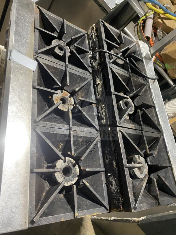 Commercial stainless steel 6-burner gas range in used condition, showing rust and wear. On legs for stability.