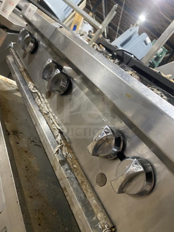 Stainless steel gas-powered 6-burner range on legs. Features control knobs, industrial grade, used condition, visible grime.