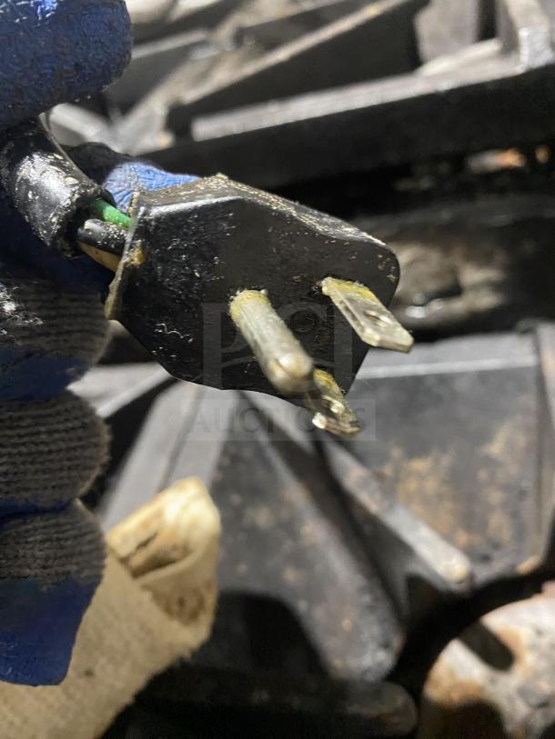 A close-up of a worn electrical plug with visible damage on one prong, held by a gloved hand.