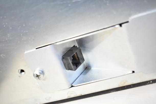 Close-up view of a cash drawer connector port on a metal surface, showing slight wear and a visible RJ11 socket.