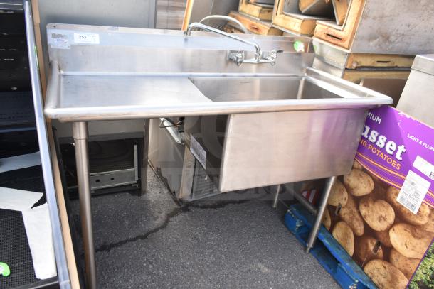 Stainless steel single bay sink with faucet and handles, used condition, visible vendor labels, on sturdy legs.