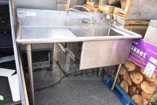 Stainless steel single bay sink with faucet and handles, used condition, visible vendor labels, on sturdy legs.