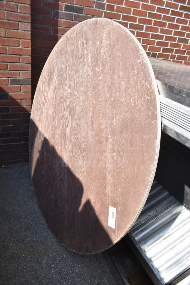 Round folding table, wood finish, leaning against a brick wall. Visible scuffs, auction tag number 266.