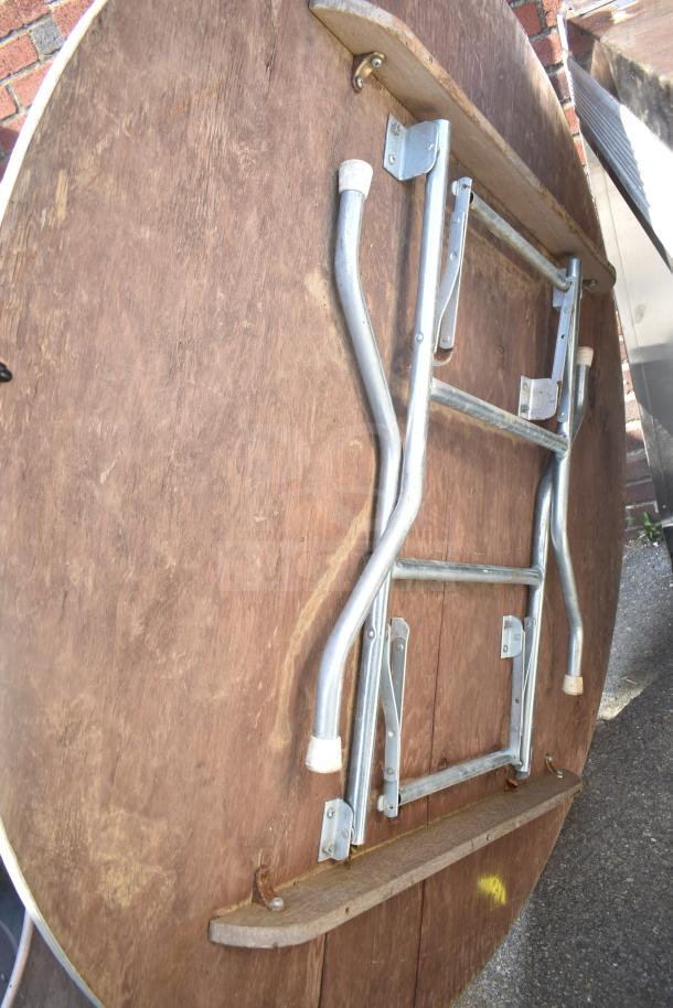 Round folding table with wooden top and metal legs. Features folding mechanism and rubber-capped feet. Used condition.