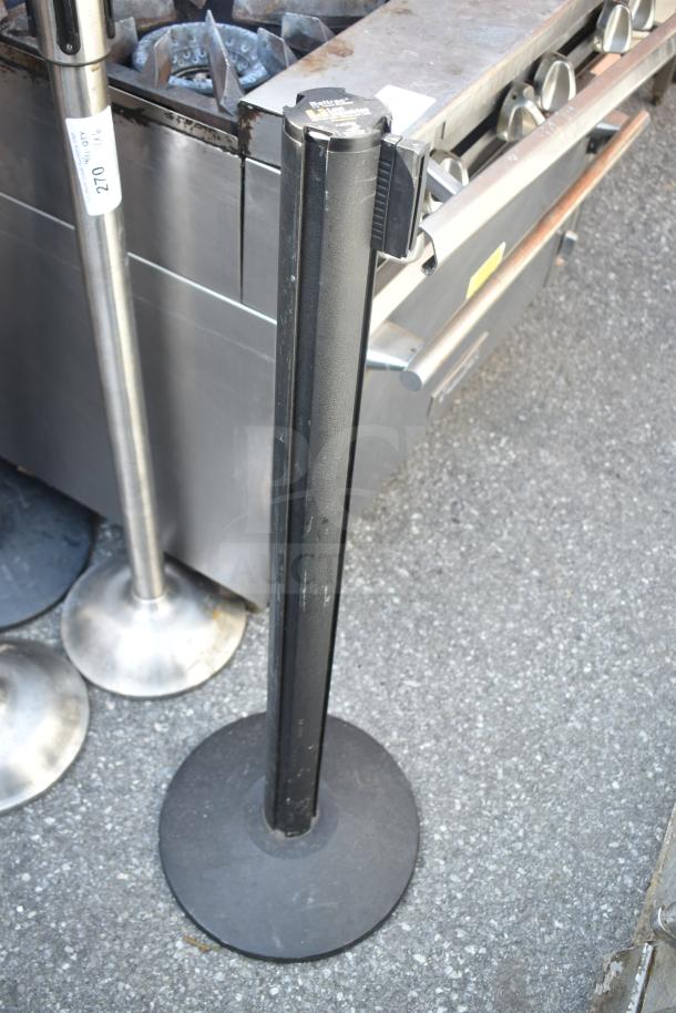 Black stanchion with retractable belt, metal base, and visible wear marks; auction includes six stanchions.