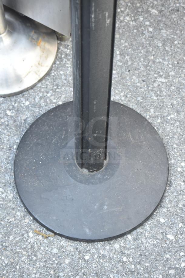 Black stanchion with a round base on pavement. Used condition, minor scratches. Part of a set of 6 available.