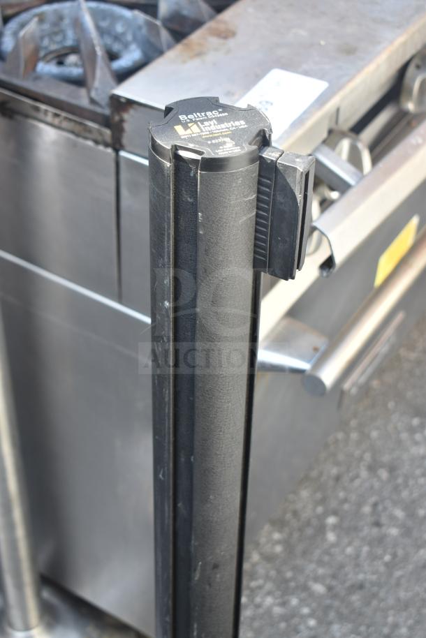 Six black Beltrac stanchions, visible logo, used condition, suitable for crowd control or queue management.