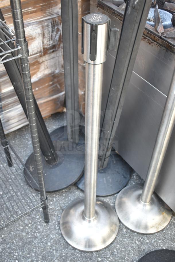 Six stainless steel stanchions with round bases, marked "BSI," indicating brand. Good condition; ideal for crowd control.