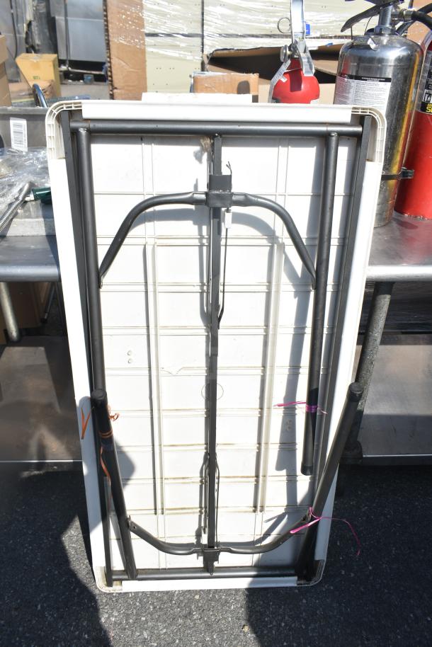 White folding table with metal legs, folded position. Slight wear visible; pink and orange ties secure the legs.