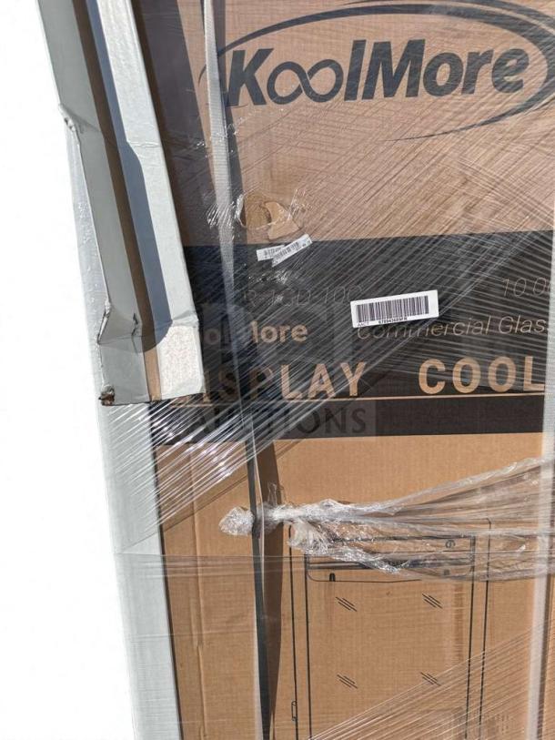 Brand new KoolMore commercial display refrigerator, model MDR1GD12C, in packaging with visible scratches and dents.